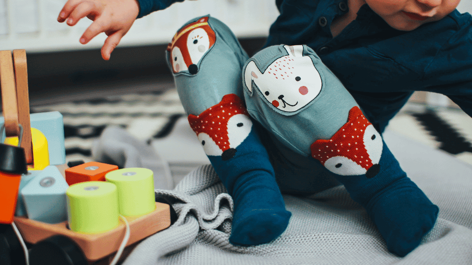 Un enfant portant des leggings avec des motifs d'animaux sur les genoux joue avec des blocs de bois colorés et des bobines sur une couverture grise, jouant avec des jeux Montessori sur un chariot pour enfant